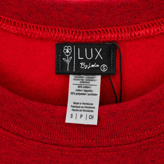 Close-up of a red knit garment showing the inner neck label, which reads LUX by Leslie, alongside a white care instruction tag.