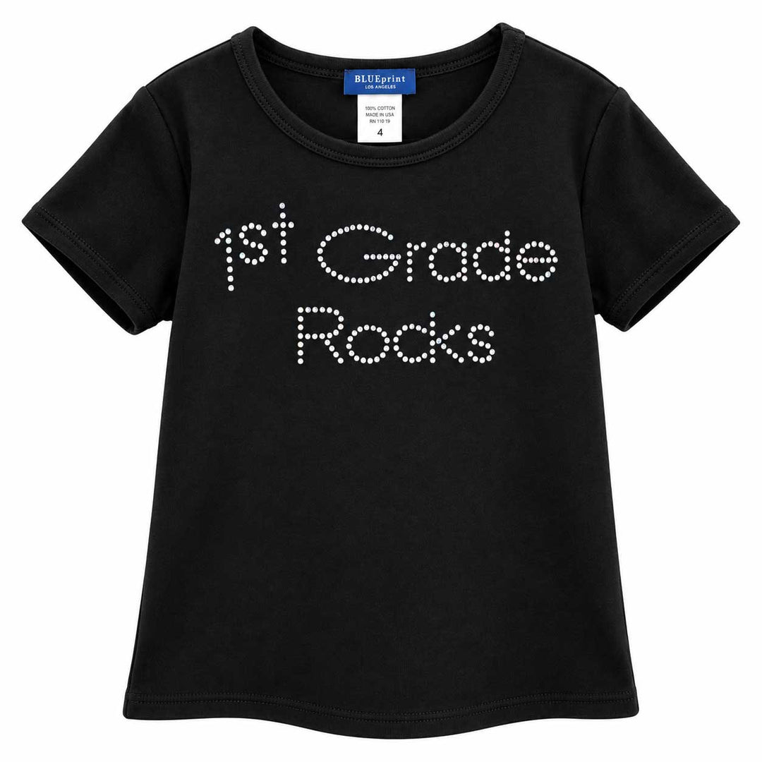 A black short-sleeve kids' t-shirt with '1st Grade Rocks' printed on the front in white rhinestone-style lettering, shown against a plain white background.
