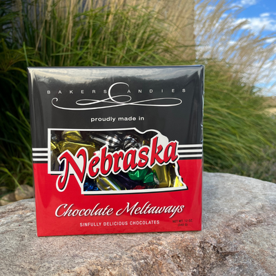 Baker's Candies 12oz Chocolate Meltaways, Nebraska Themed with Nebraska cut out to show the product. Product is placed on a rock with a grassy background.