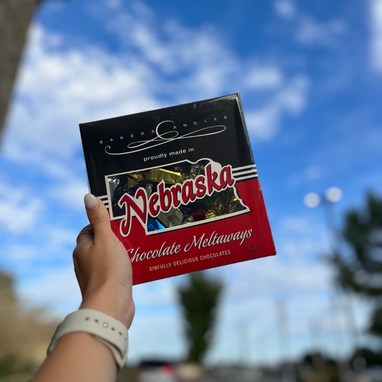 Baker's Candies 12oz Chocolate Meltaways, Nebraska Themed with Nebraska cut out to show the product being held by a hand on a blurred background.