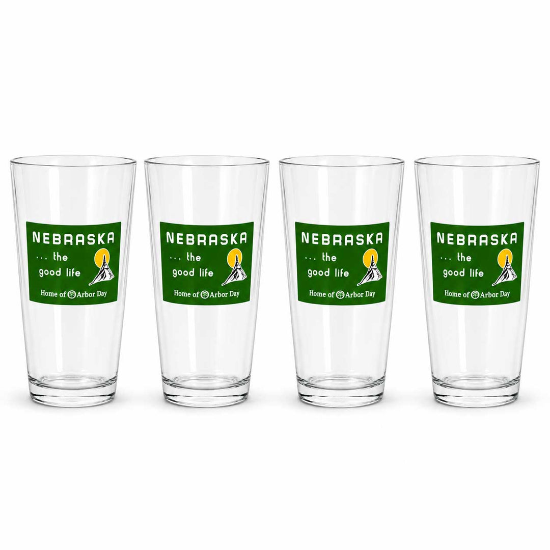 Four clear drinking glasses arranged in a row on a white background, each printed with a green Nebraska graphic that reads 'the good life' and 'Home of the Nebraska Cornhuskers.'.