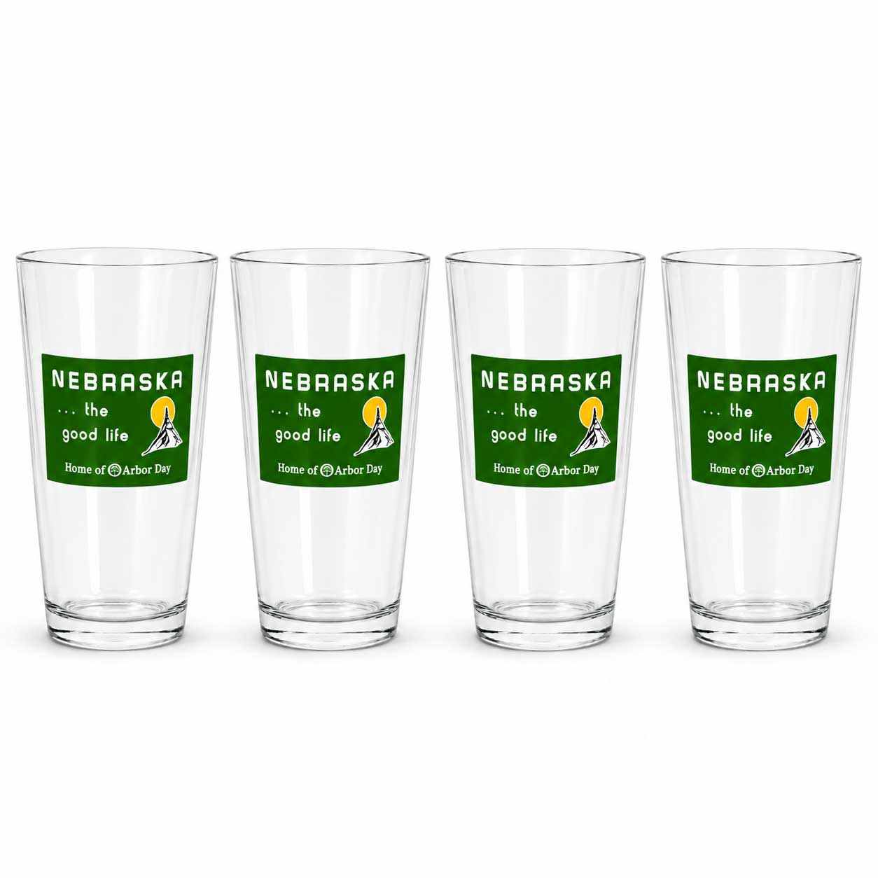 Four clear drinking glasses arranged in a row on a white background, each printed with a green Nebraska graphic that reads 'the good life' and 'Home of the Nebraska Cornhuskers.'.