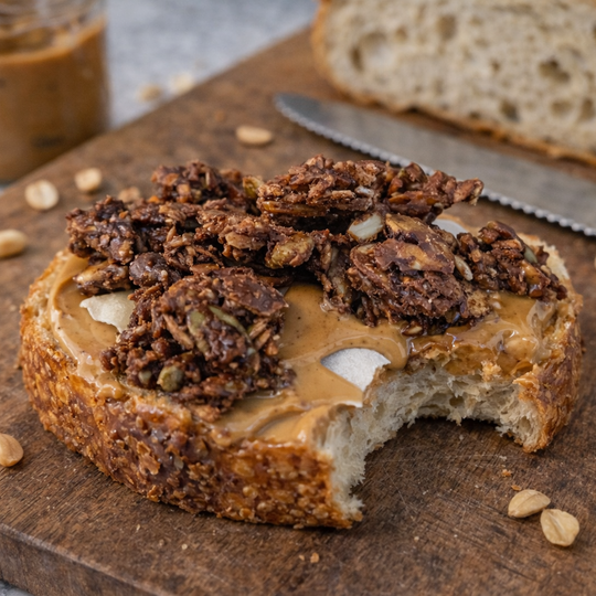 Slice of bread with peanut butter and granola on a wooden cutting board, with a jar of peanut butter and knife in the background.