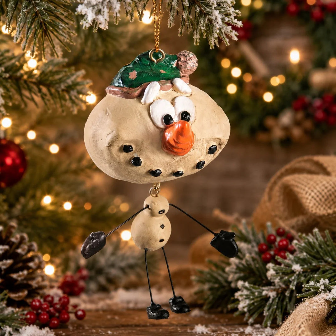 A handmade clay Christmas snowman ornament hanging from a gold loop on a decorated Christmas tree. The snowman has a beige body, black stick-like arms and legs, a green hat, and a bright orange carrot nose, set against a warm, blurred holiday background with lights and pine branches.
