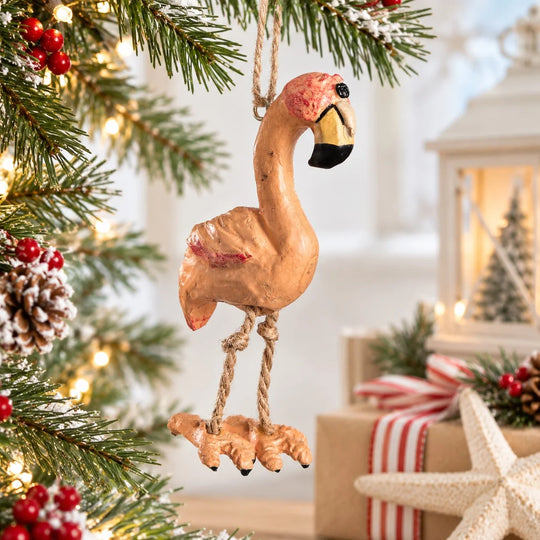 A rustic wooden flamingo Christmas ornament hanging from a pine tree branch with warm holiday lights, wrapped in a cozy festive setting with wrapped gifts and a lantern in the background.