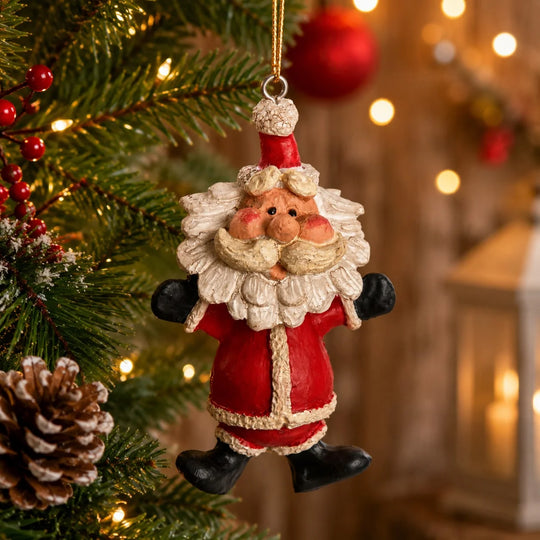 A hanging Christmas ornament shaped like a Santa Claus figure, shown attached to a real Christmas tree with warm string lights and a softly blurred indoor holiday background.