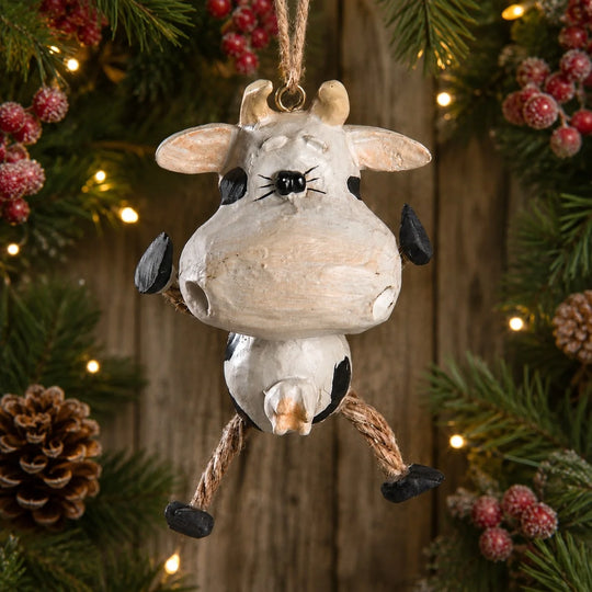 A rustic Christmas hanging ornament shaped like a cow, with a distressed off-white body, black spots, small horns, and rope legs, displayed against a festive holiday background of pine branches, berries, and warm string lights.
