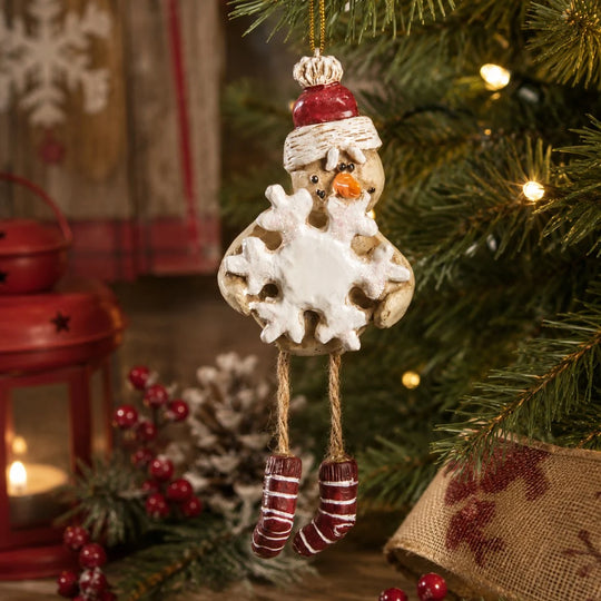 A rustic snowman Christmas ornament hanging from a decorated tree. The ornament features a weathered wood body with a white snowflake-shaped belly, a red and white knit hat, and red-and-white striped socks.