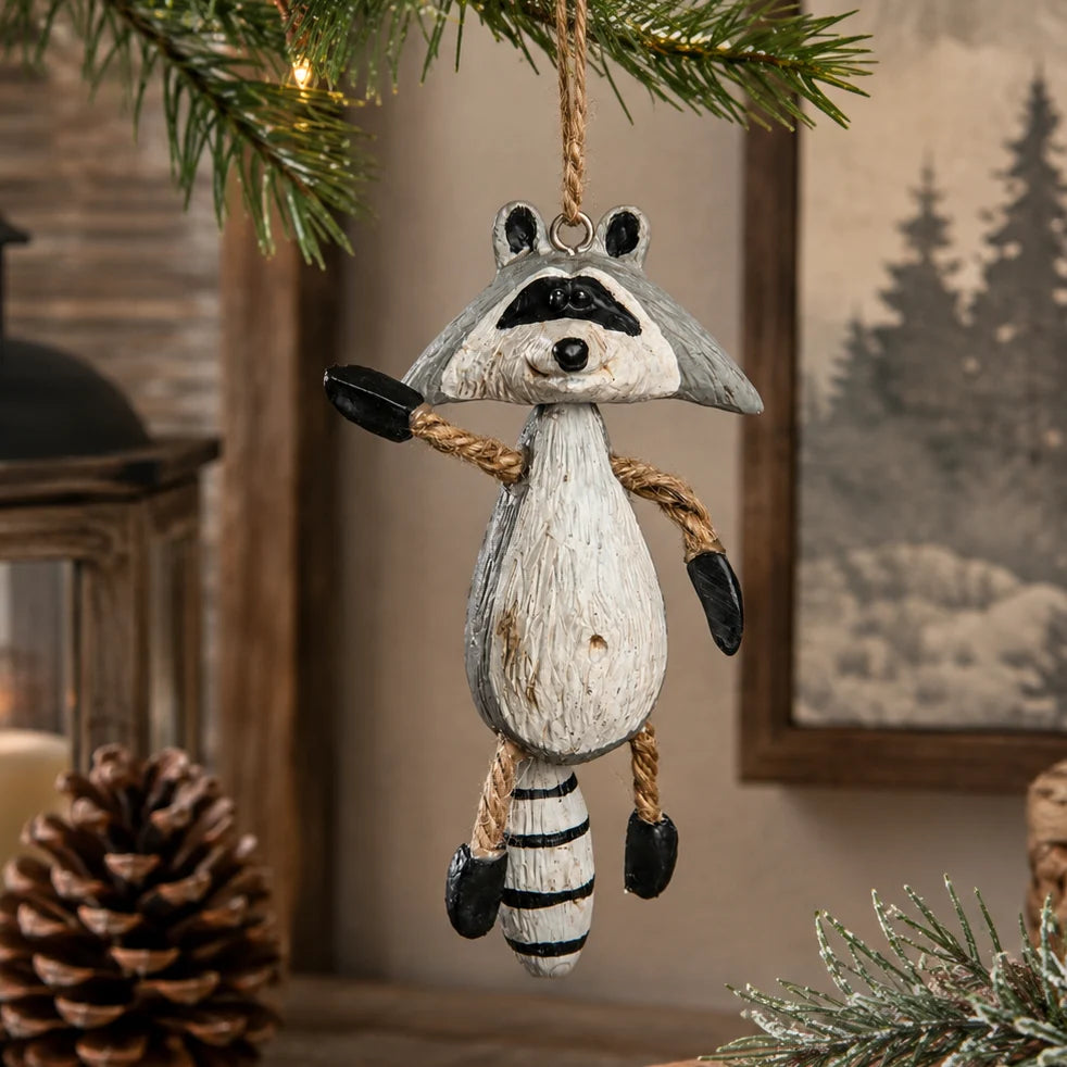 A hanging raccoon Christmas ornament made to look like a distressed wood carving, suspended from a pine branch in a rustic indoor setting with a pinecone and framed picture in the background.
