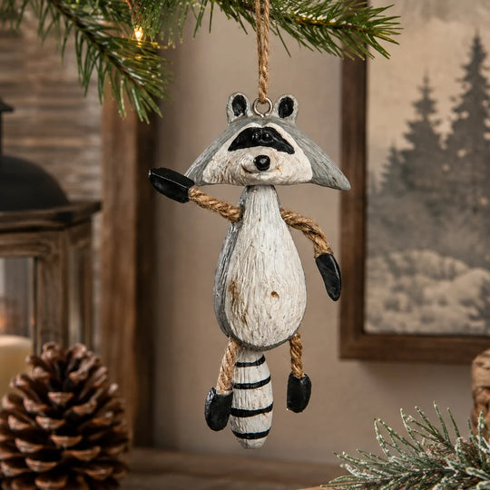 A hanging raccoon Christmas ornament made to look like a distressed wood carving, suspended from a pine branch in a rustic indoor setting with a pinecone and framed picture in the background.