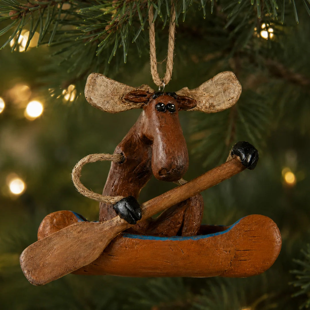 A rustic moose-shaped Christmas ornament sitting in a canoe, hanging from a tree branch with warm bokeh lights in the background.