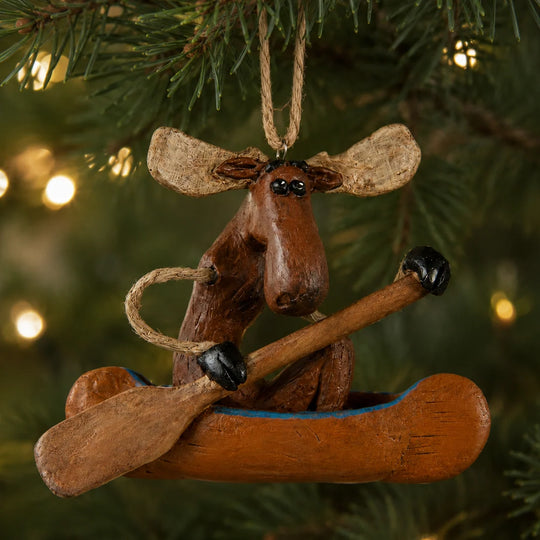 A rustic moose-shaped Christmas ornament sitting in a canoe, hanging from a tree branch with warm bokeh lights in the background.