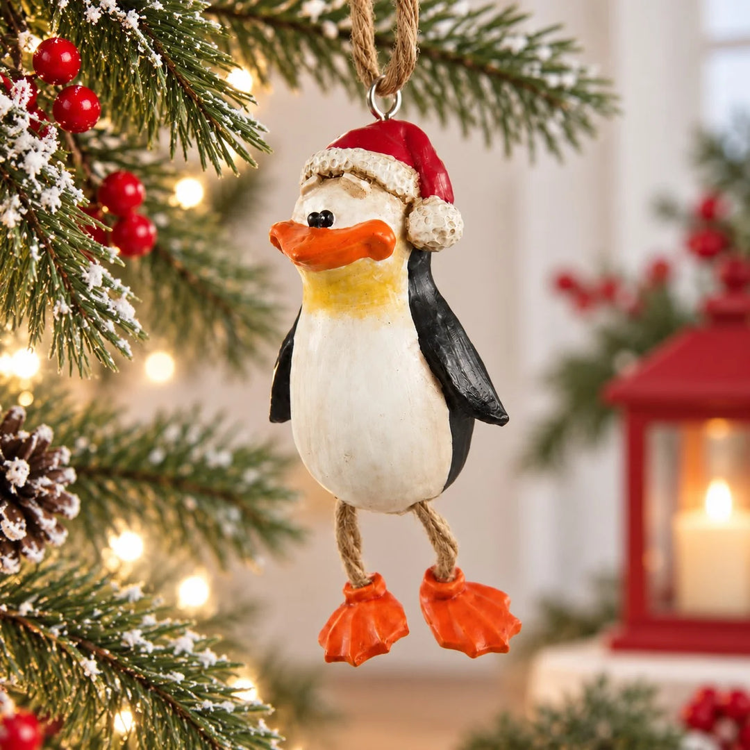 A Christmas penguin ornament hanging from a tree branch. The ornament is painted white on the front with black wings and feet, an orange beak and feet, and a red Santa hat with white trim.