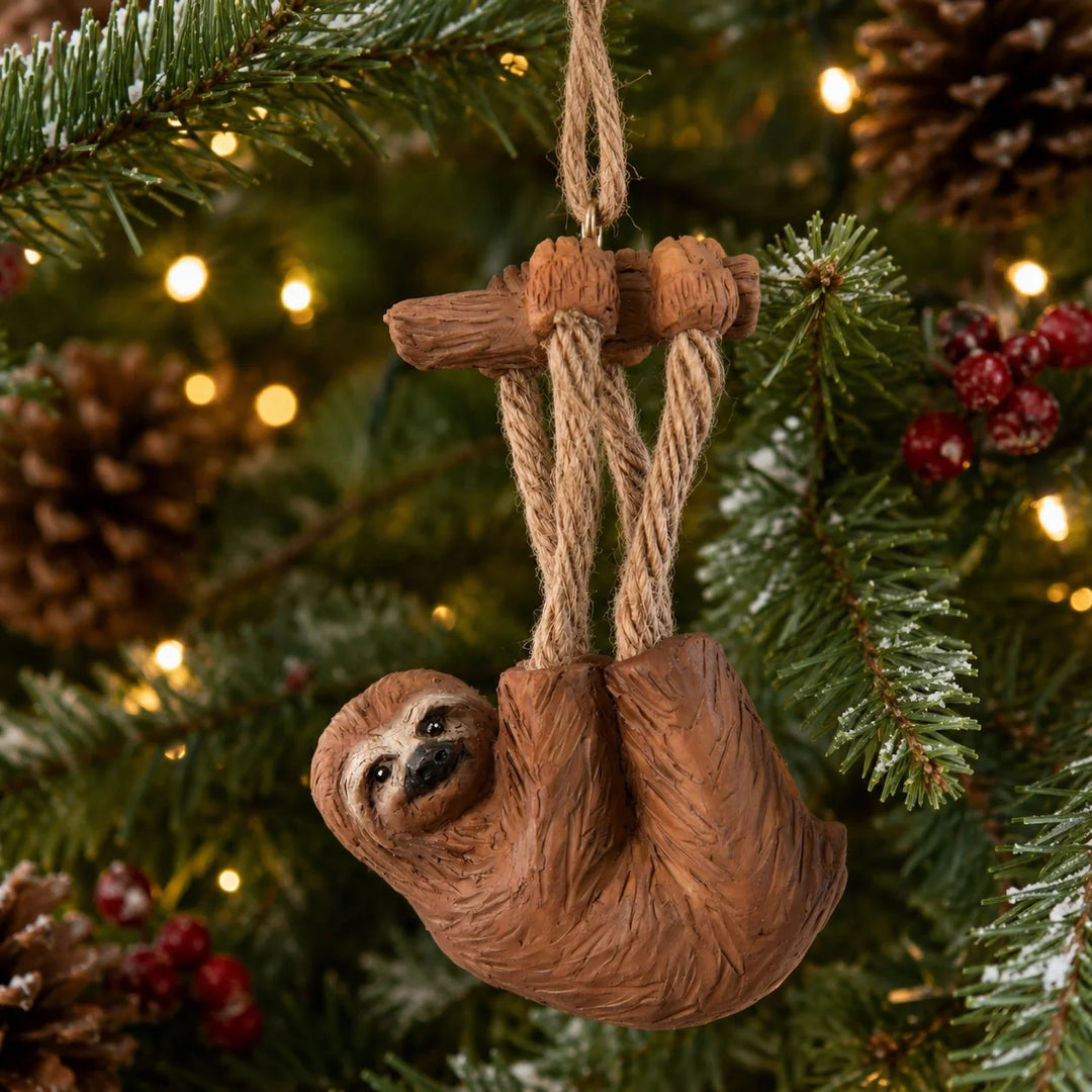 A sloth-shaped Christmas ornament hanging from a rope and branch, displayed on a decorated Christmas tree with pinecones and warm string lights in the background.