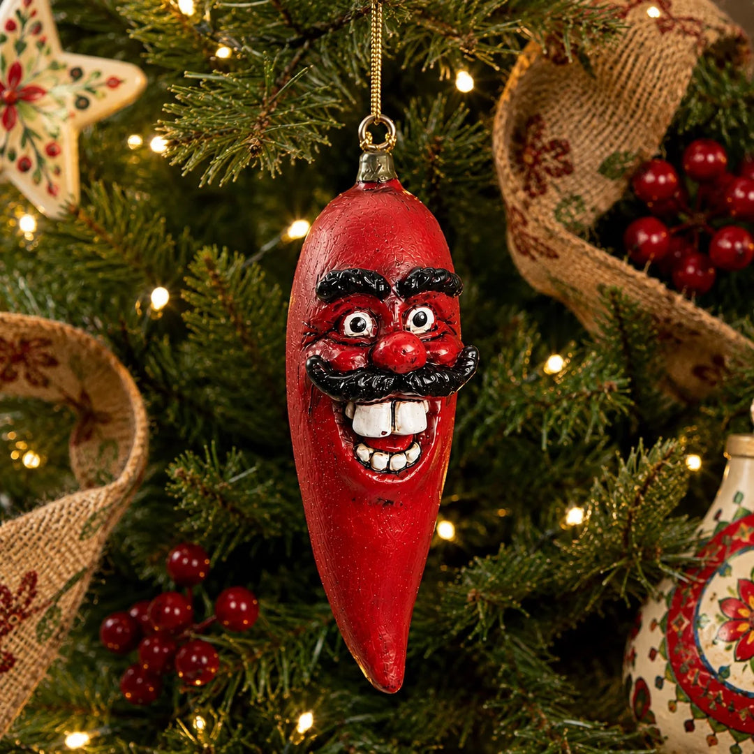A red chili pepper Christmas ornament with a smiling face and black mustache hanging on a decorated Christmas tree with lights and burlap garlands.