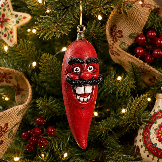 A red chili pepper Christmas ornament with a smiling face and black mustache hanging on a decorated Christmas tree with lights and burlap garlands.