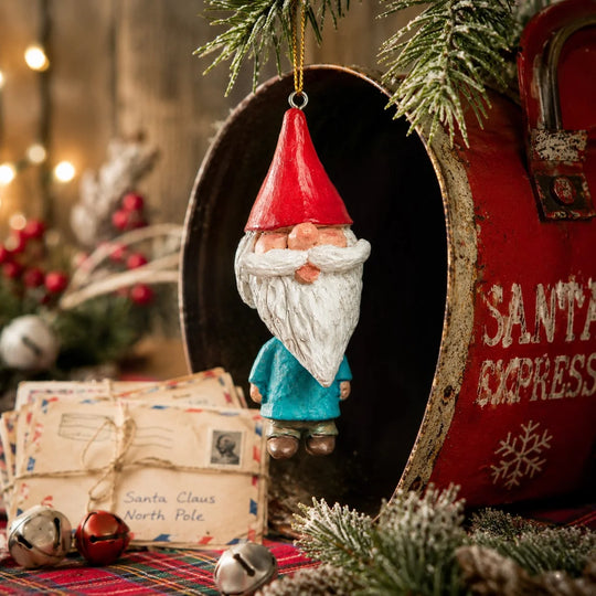 A Christmas gnome ornament with a red hat, white beard, and blue outfit hangs in front of an open red Santa Express mailbox decorated with pine branches, wrapped letters, and small bells.