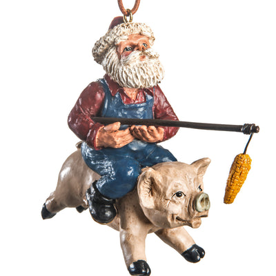 Santa-farmer ornament, wearing Santa hat, red collared shirt, bib overalls and holding an ear of corn on the end of a long stick and riding a pig, shown on a white background