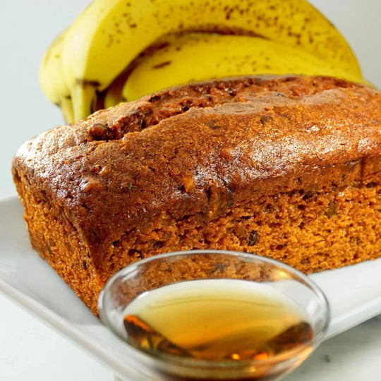 Loaf of banana bread on a white plate with bananas and a small bowl of syrup in the background.