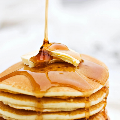 A real-life photo of Buttermilk Pancakes Stacked with butter and syrup being poured on top.
