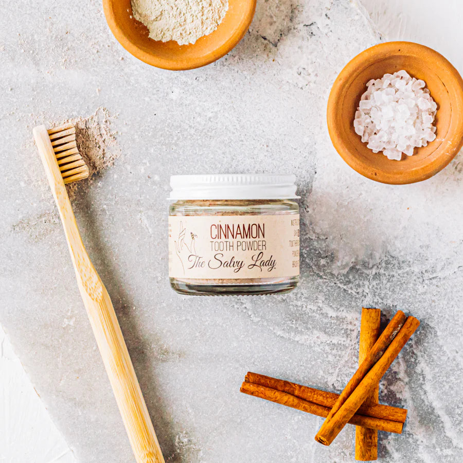 Jar of cinnamon tooth powder with a bamboo toothbrush and cinnamon sticks on a textured surface.