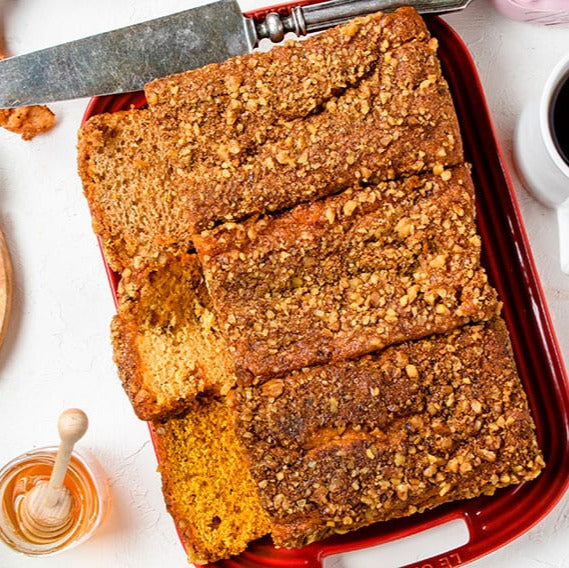Loaf of bread in a red pan with a knife, surrounded by grapes, cookies, and a cup of coffee on a white surface.