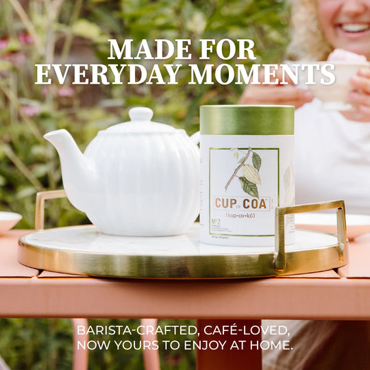 A white teapot and a cylindrical canister labeled CUP.COA sit on a round marble tray with gold-toned handles, placed on a wooden table outdoors.