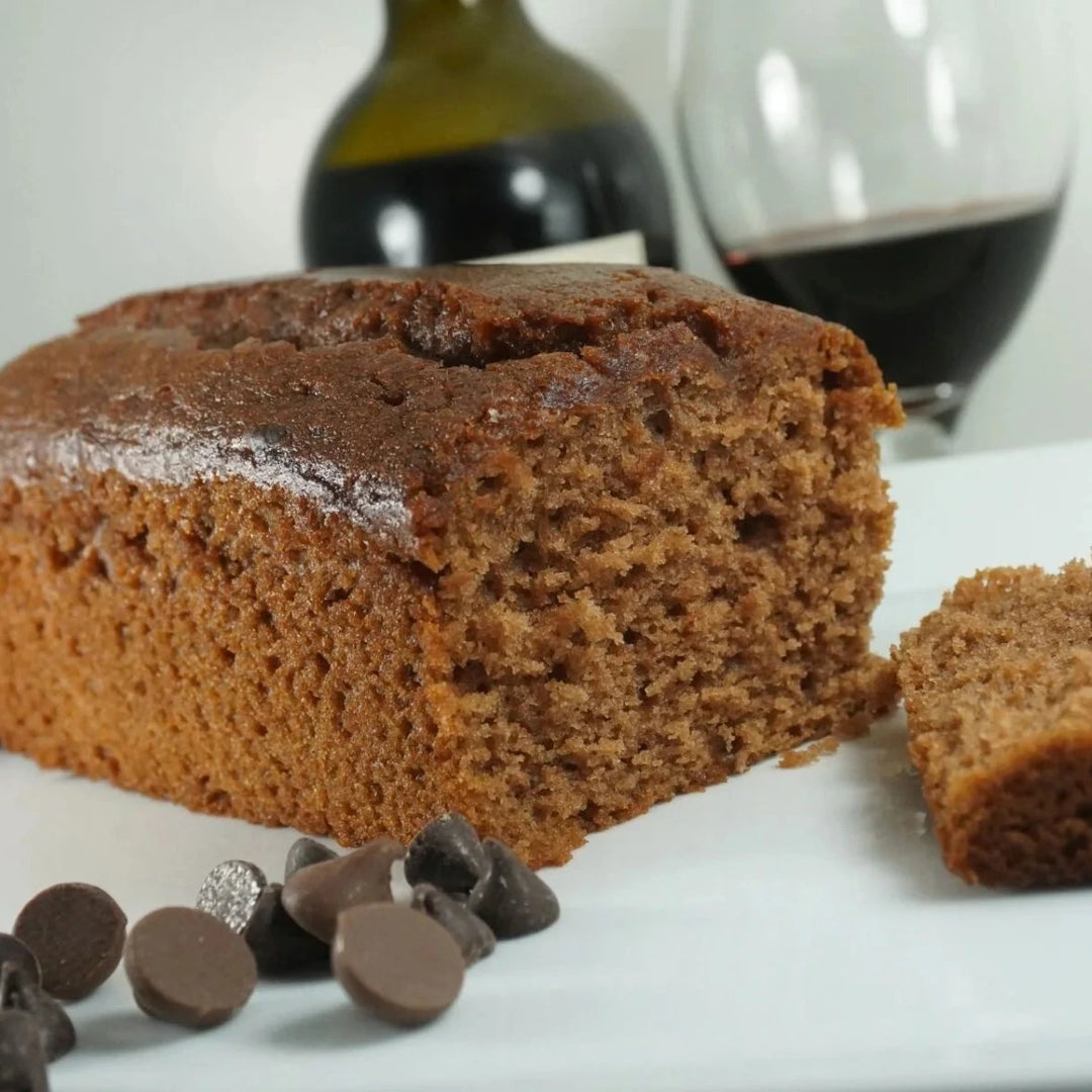 Chocolate merlot loaf cake with a slice cut, surrounded by chocolate chips on a white surface.