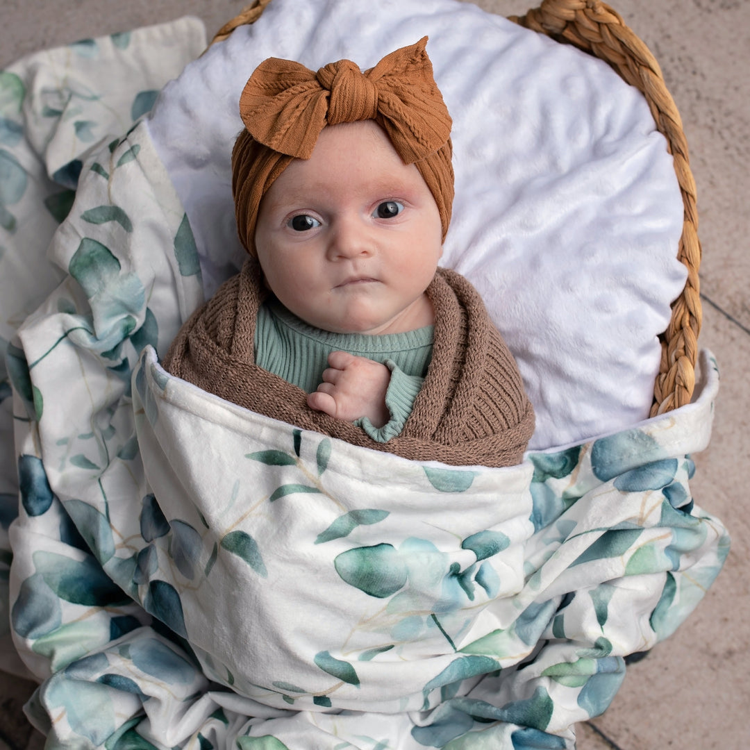Newborn baby wrapped in a floral blanket with a headband, sitting in a woven basket.