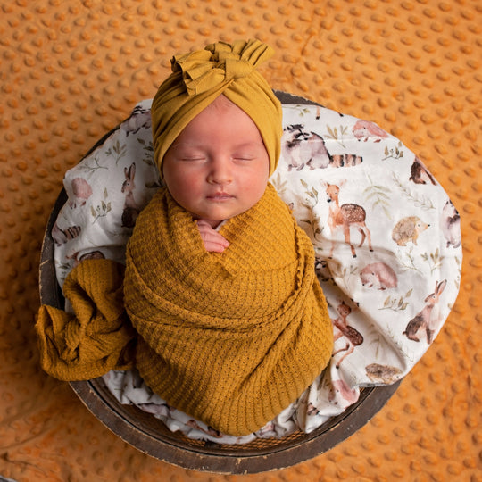 Newborn baby wrapped in mustard yellow fabric with a matching headband, lying on a patterned blanket.