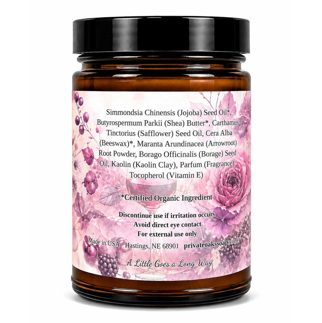 An amber glass jar of seed oil with a black lid and a pink floral label, photographed on a clean white background. The label lists ingredients such as Simmondsia Chinensis seed oil and butyrospermum parkii (shea) butter.