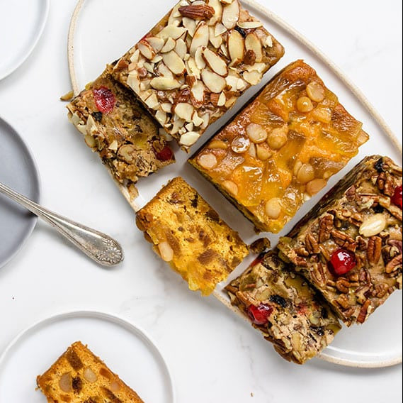 Slices of fruitcake with nuts and cherries on a white surface with plates and a fork.