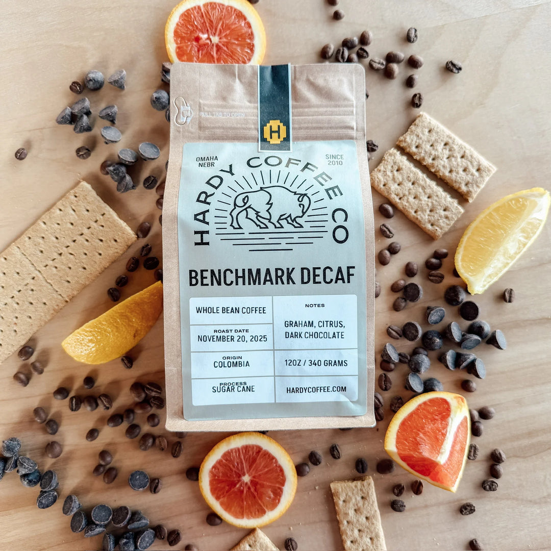 Hardy Coffee Co. Benchmark Decaf package surrounded by coffee beans, orange slices, and crackers on a wooden surface.