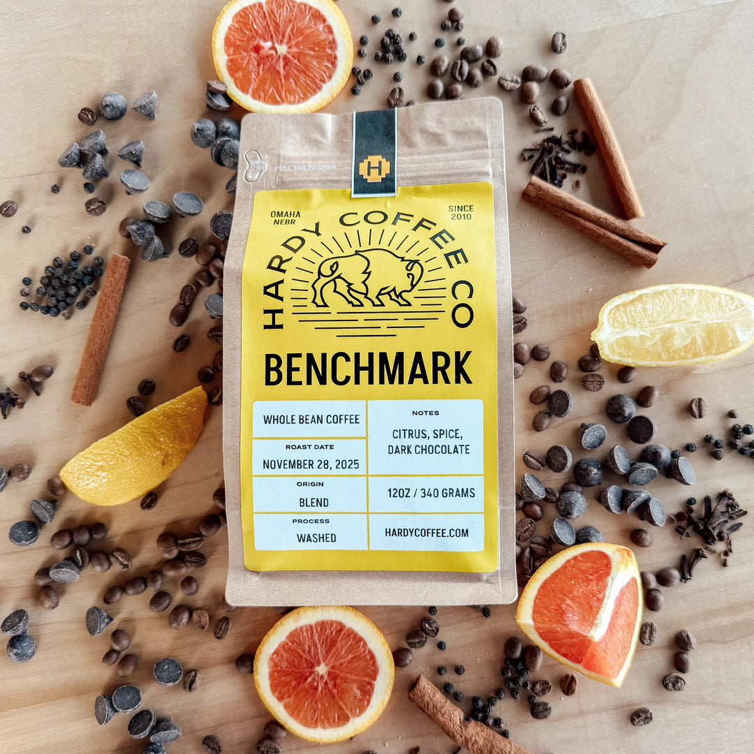 Hardy Coffee Co Benchmark coffee package surrounded by coffee beans, citrus slices, and cinnamon on a wooden surface.