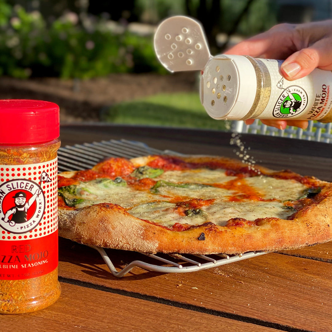 Person seasoning a pizza with a bottle of pizza seasoning on a wooden table outdoors.