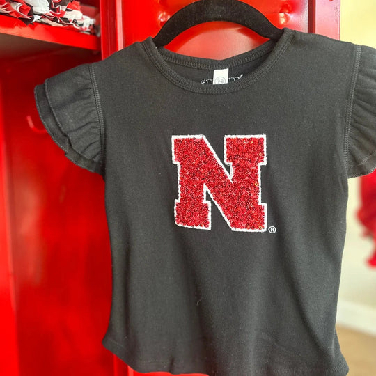 A black short-sleeve t-shirt with ruffled sleeves hangs on a black hanger in front of a red locker. The shirt features a large red glittery 'N' logo outlined in white on the front.
