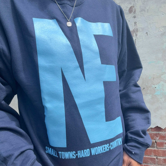 A close-up of a person wearing a navy blue sweatshirt with a large light blue 'NE' graphic and the text 'SMALL TOWNS-HARD WORKERS-COUNTRY' printed along the bottom hem.