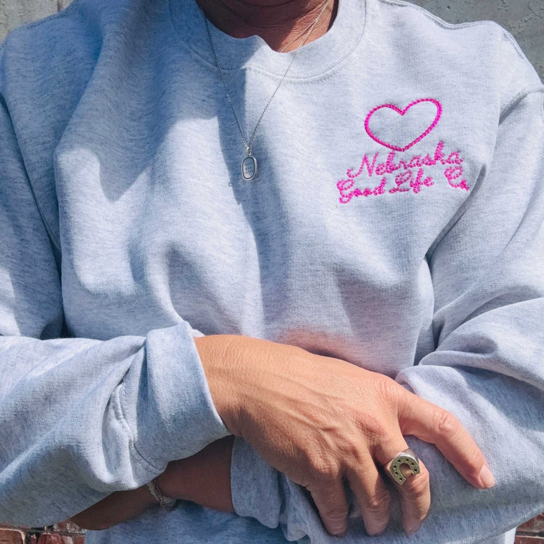 A close-up of a person wearing a light heather gray sweatshirt with a pink embroidered heart and the words 'Nebraska Good Life Co' on the left chest.
