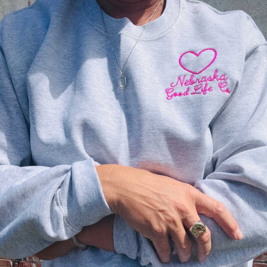 A close-up of a person wearing a light heather gray sweatshirt with a pink embroidered heart and the words 'Nebraska Good Life Co' on the left chest.