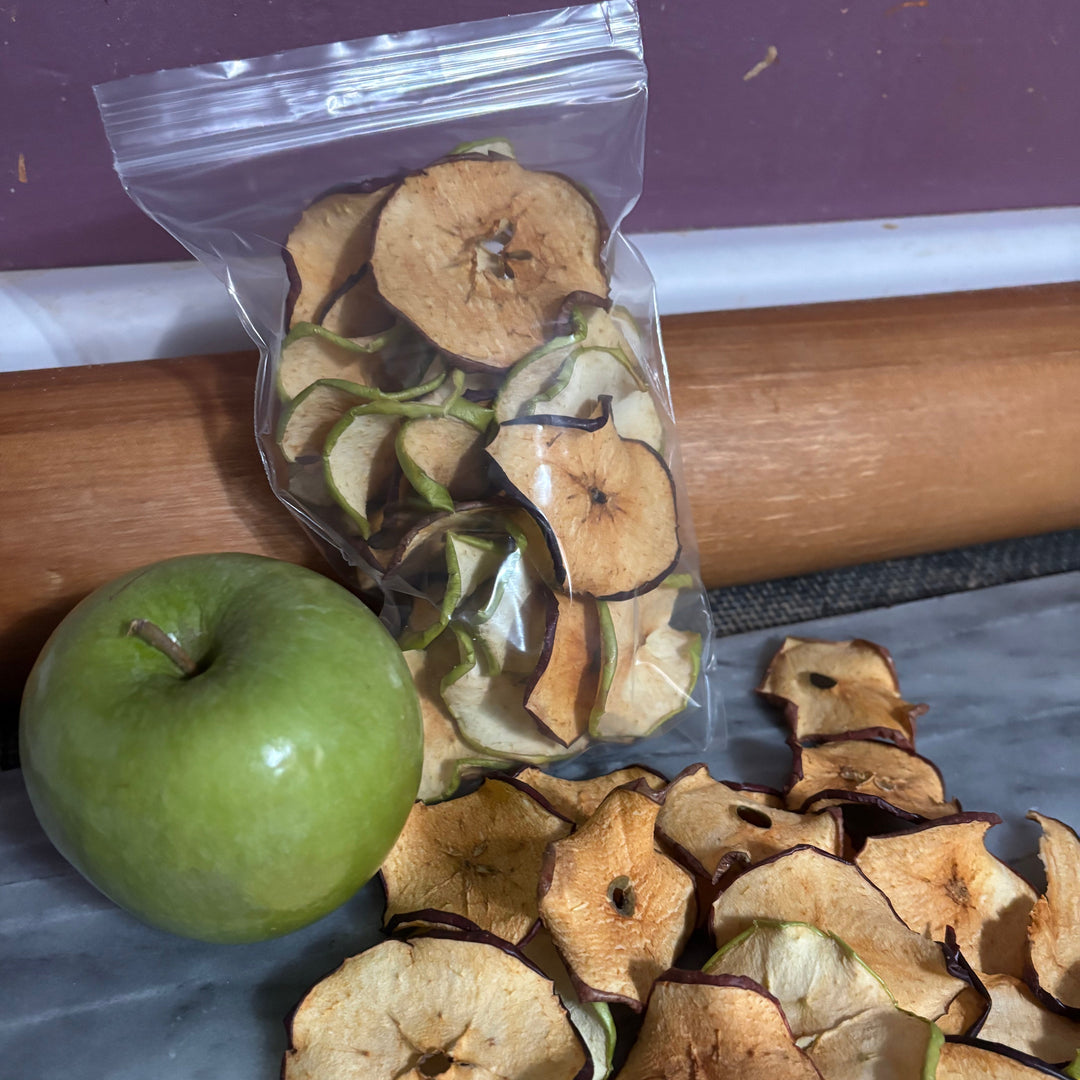 Dried apple slices in a clear bag next to a green apple on a wooden surface.