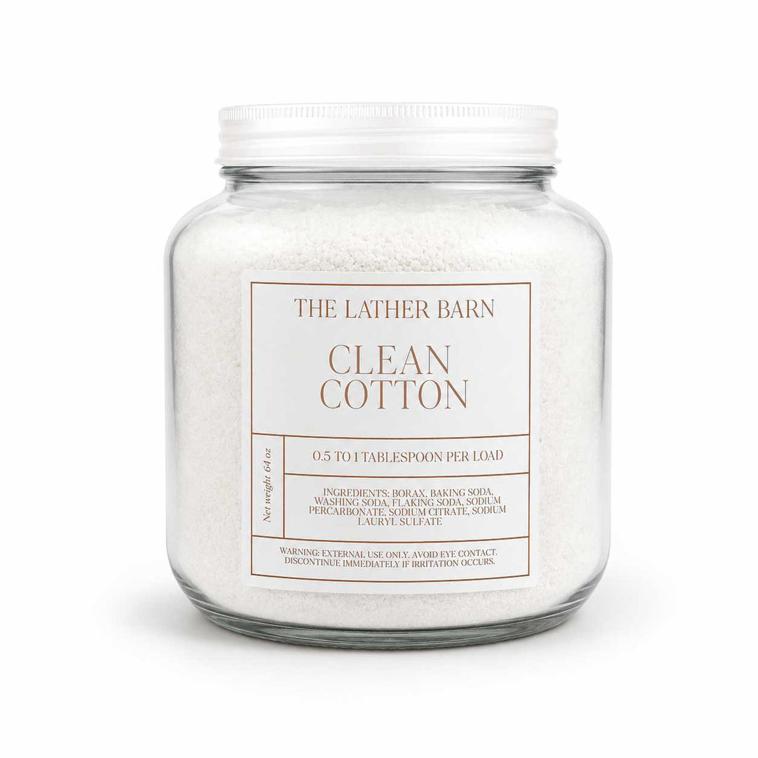 A glass jar filled with white cotton laundry detergent, featuring a white metal lid and a white label that reads 'THE LATHER BARN CLEAN COTTON'.