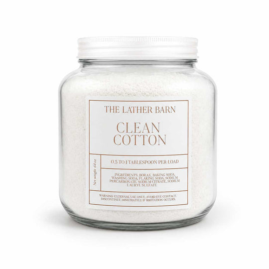 A glass jar filled with white cotton laundry detergent, featuring a white metal lid and a white label that reads 'THE LATHER BARN CLEAN COTTON'.