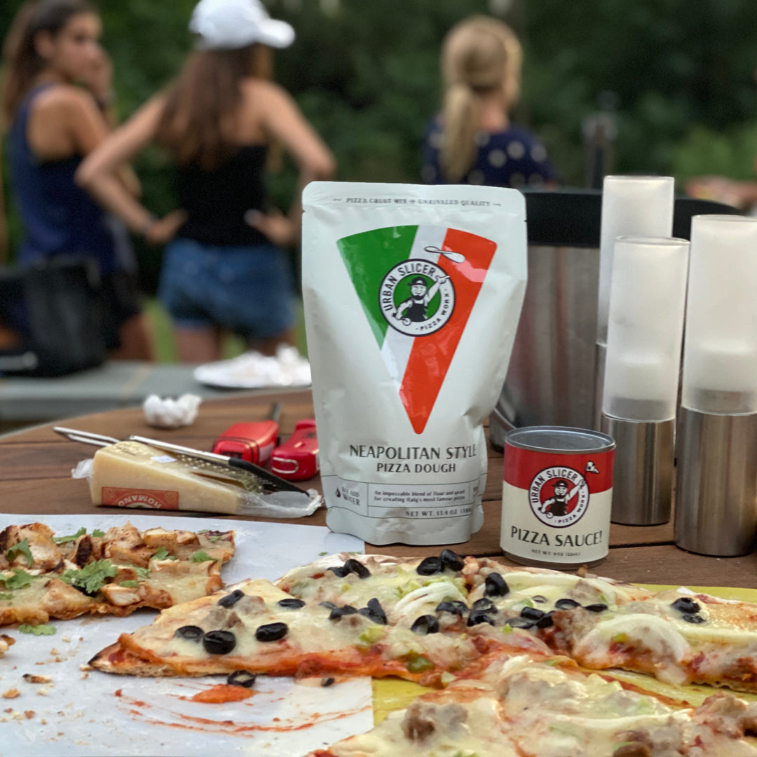 Pizza-making setup with pizza dough, sauce, and ingredients on a table outdoors.