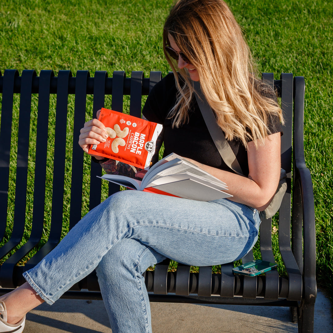 Woman sitting on a bench reading a book and eating cashews outdoors.