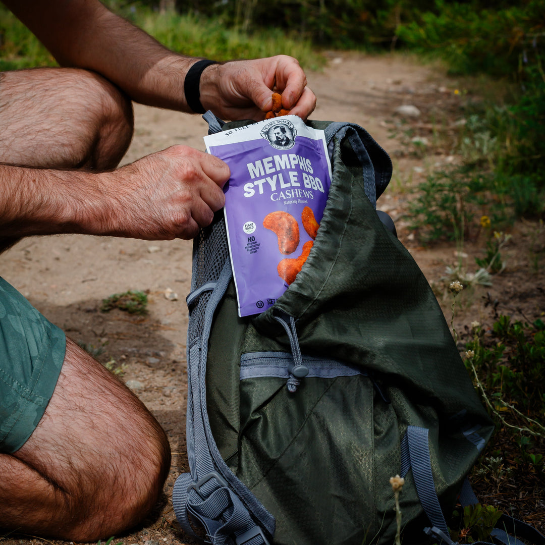 Person outdoors with a green backpack and a package of Memphis cashews on a dirt path.