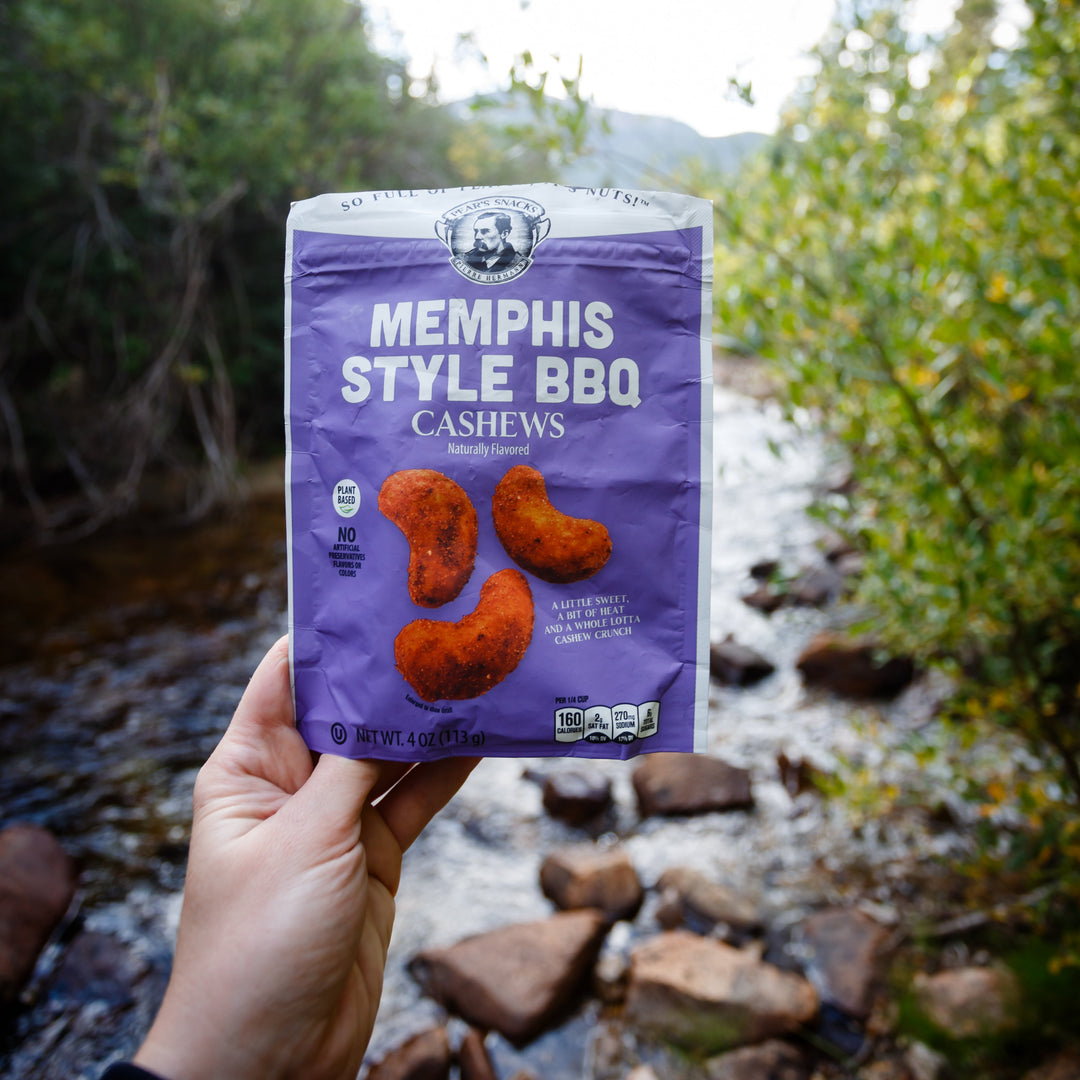 Hand holding a package of Memphis Style BBQ Cashews against a natural background with trees and water.