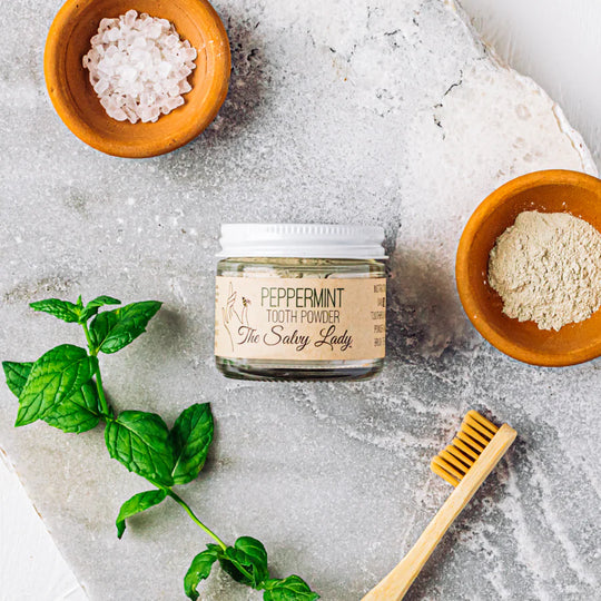 Jar of peppermint tooth powder with a wooden toothbrush and bowls of ingredients on a textured surface