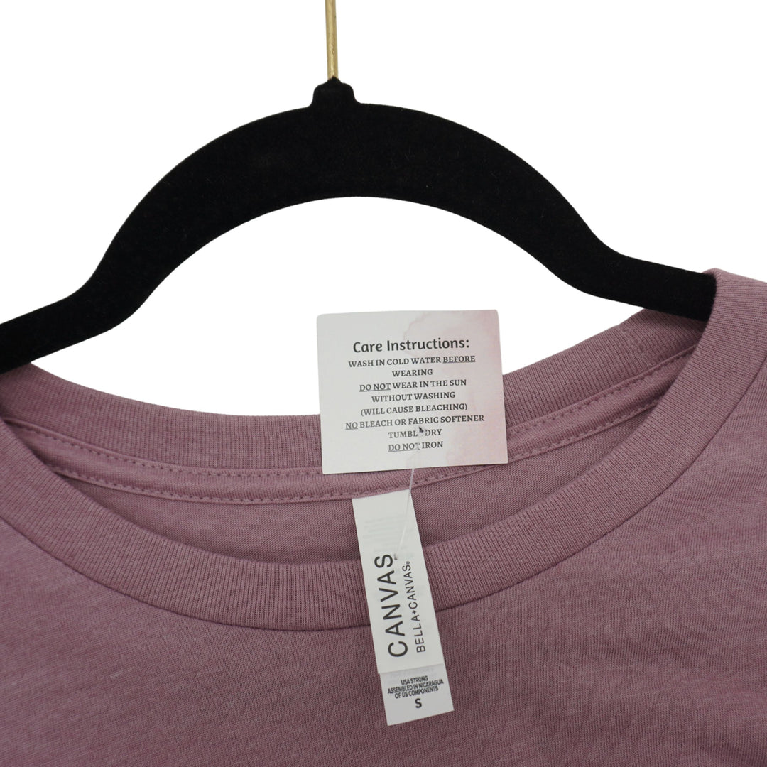 Close-up of a mauve-pink crewneck t-shirt hanging on a black hanger, showing the inner neck labels for 'CANVAS' by Bella+Canvas.