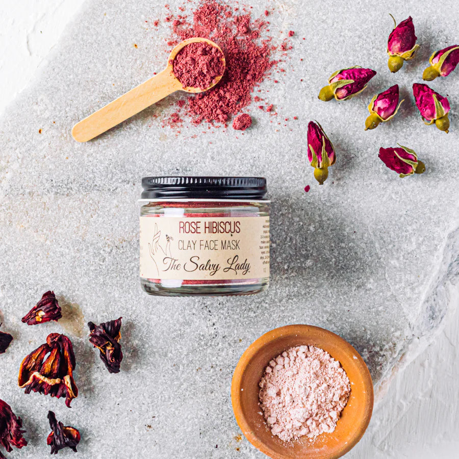 Jar of Rose Hibiscus Clay Face Mask with dried flowers and a wooden spoon on a light background