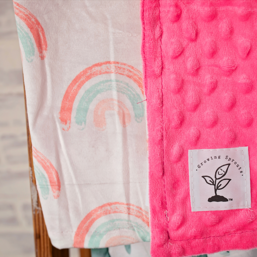 Pink textured blanket with a label and a white blanket with rainbow design on a wooden surface.